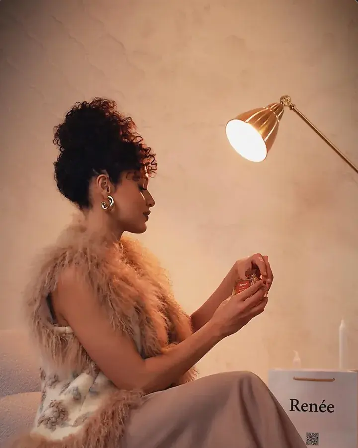 A woman in a fur stole holding a perfume bottle under a lamp